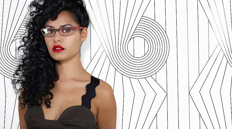 Eine Frau steht vor einer Wand mit schwarzem Linien Design und trägt eine rote Brillenfassung für Damen