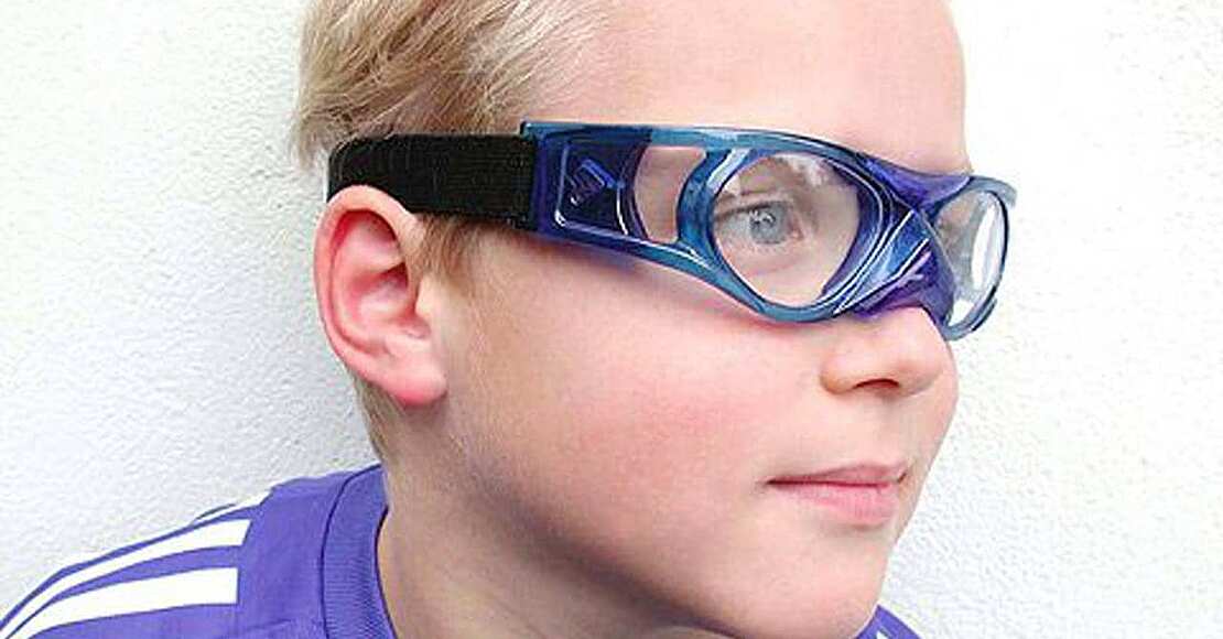 Ein Junge steht vor einer weißen Wand und trägt eine blau/schwarze Kindersportbrille für den Sportunterricht