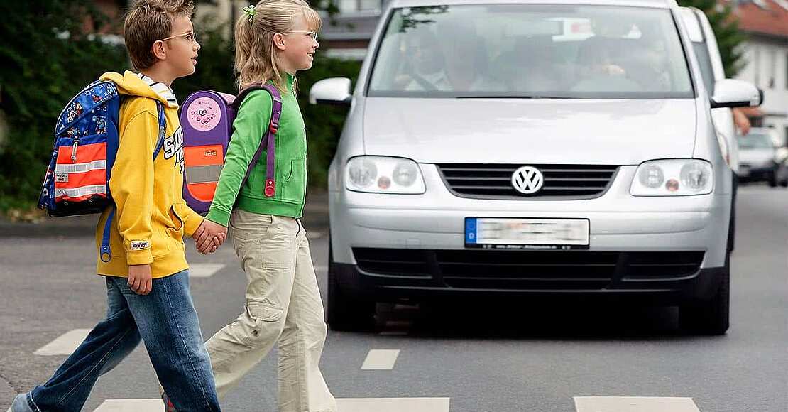 Zwei Schulkinder überqueren einen Zebrastreifen, an dem Autos warten und hiermit Sicherheit für die Kinder im Straßenverkehr bieten